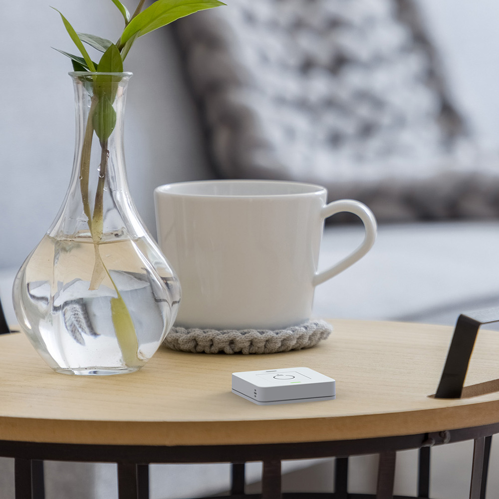 Weißer, quadratischer myStrom WiFi Button Plus auf Tisch neben einer Vase mit einer Pflanze und einer Kaffeetasse