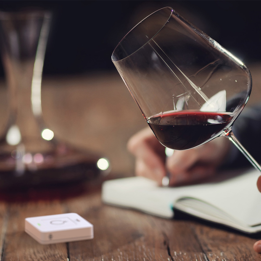 Weißer, quadratischer myStrom WiFi Button Plus auf einem Tisch neben einer Frau, die ein Buch liest und einen Wein trinkt